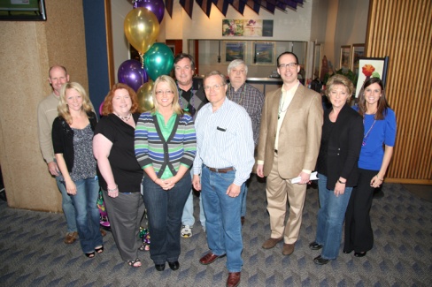 A group of ten people standing in a room with a blue carpeted floor and a wooden wall in the background. All are smiling and posing for the photo. The group appears to be of different ages and ethnicities and they are standing close together in a group.<br /><br />Behind the group there are several colorful balloons in shades of purple green and gold and a number of framed pictures hanging on the wall. The overall mood of the photo is happy and celebratory.