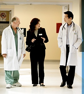 Three medical professionals standing in a hospital corridor. They are all wearing white lab coats and stethoscopes around their necks. The man on the left is an older man with white hair wearing a green scrub suit and a stethoscope around his neck. The woman in the middle is a middle-aged woman with dark hair and a black blazer. She is holding a clipboard and appears to be engaged in a conversation with the two men on either side of her. The background shows a door and a sign on the wall. The overall mood of the image is serious and professional.