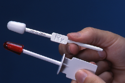 A hand holding two syringes. The syringe on the left is white with a red cap and a white handle. The handle has the letters "RX" written on it in black. The needle on the right is also white and appears to be made of plastic. The background is a solid blue color.