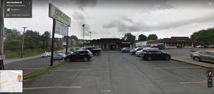 A street view of a parking lot with several cars parked in it. The parking lot is located on the right side of the image with a building on the left side. The building appears to be a convenience store or a restaurant as there is a sign that reads "McDonald's" above the entrance. The sky is cloudy and there are trees and bushes in the background. The road is empty with no cars or people visible. There is a map on the bottom right corner of the photo showing the location of the parking lot.