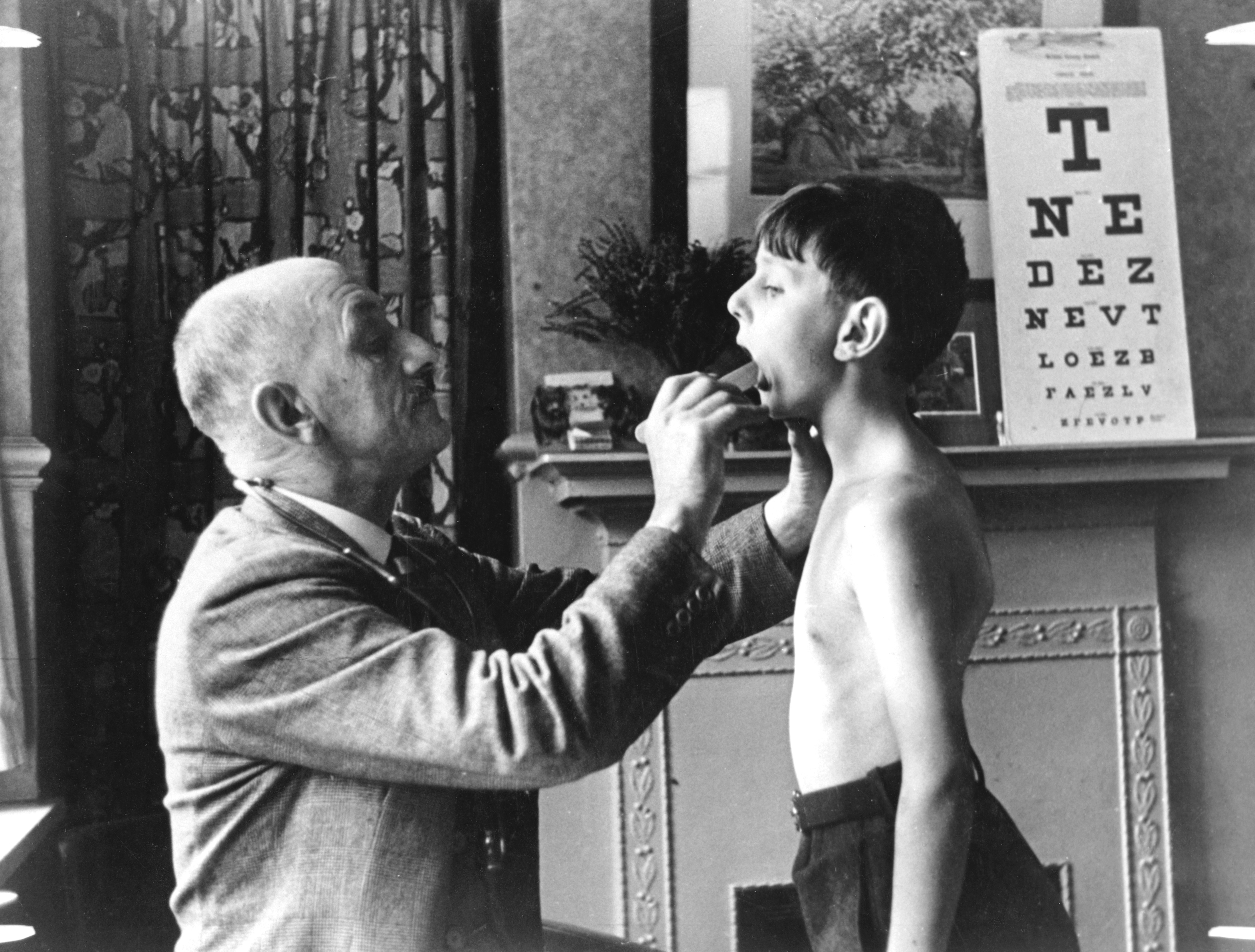 A black and white photograph of an elderly man and a young boy in a room. The man is standing on the left side of the image holding a small object in his hand and appears to be examining the boy's throat. The boy is standing in front of him with his mouth open and his eyes closed. He is shirtless and is looking up at the man with a concerned expression on his face. In the background there is a window with curtains and a sign that reads "TNE DEZ NEVIT LOEZ" which is a type of eye chart. The image appears to have been taken in the early 20th century.