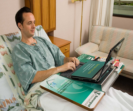 A young man sitting in a hospital bed with a laptop on a tray in front of him. He is wearing a green hospital gown and has a smile on his face. The tray is made of cardboard and has the logo of a hospital on it. The laptop is open and the screen is turned on. The man is typing on the keyboard and appears to be focused on the task at hand. In the background there is a beige armchair and a window with white curtains. The room is well-lit with natural light coming in from the window.
