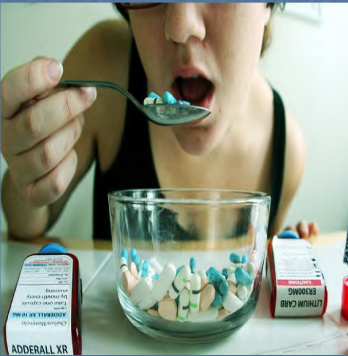 A young woman sitting at a table with a glass bowl in front of her. She is holding a spoon with blue and white pills in her mouth. The woman is wearing a black tank top and appears to be taking a bite out of the spoon. On the table there are several other pills and capsules scattered around the bowl. The background is blurred but it seems like the woman is in a room with a white wall.