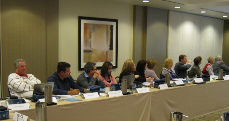 A group of people sitting at a long table in a conference room. The table is covered with a beige tablecloth and has nameplates and water bottles on it. There are several people sitting in rows of chairs facing the front of the room attentively listening to a speaker. On the wall behind the table there is a framed painting hanging. The room appears to be well-lit with natural light coming in from the windows on the right side of the image.