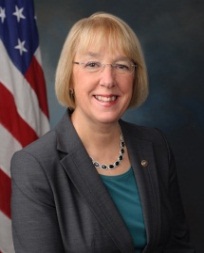 A professional headshot of a woman. She has blonde hair and is wearing glasses. She is smiling and looking directly at the camera. The woman is wearing a gray blazer over a green top and a black necklace. Behind her there is an American flag. The background is a dark blue color.