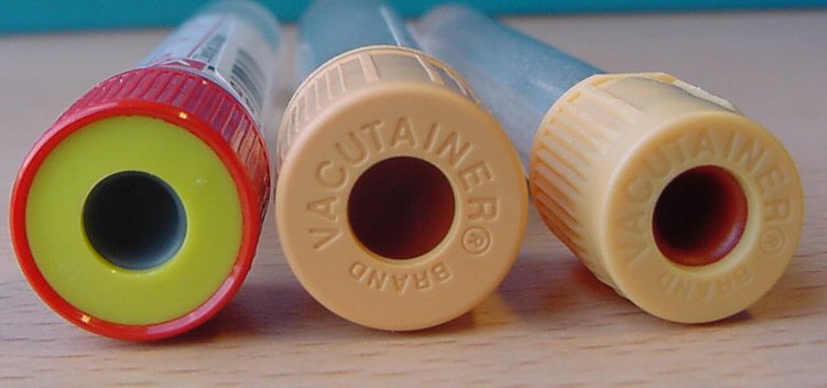 Three tubes of different colors - red yellow and blue - arranged in a row on a wooden surface. The tubes are cylindrical in shape and have the word "Vacutainer" written on them in white letters. The red tube has a small hole in the center while the yellow tube is slightly larger than the blue tube. All three tubes appear to be new and unused. The background is blurred but it appears to be a wooden table or countertop.