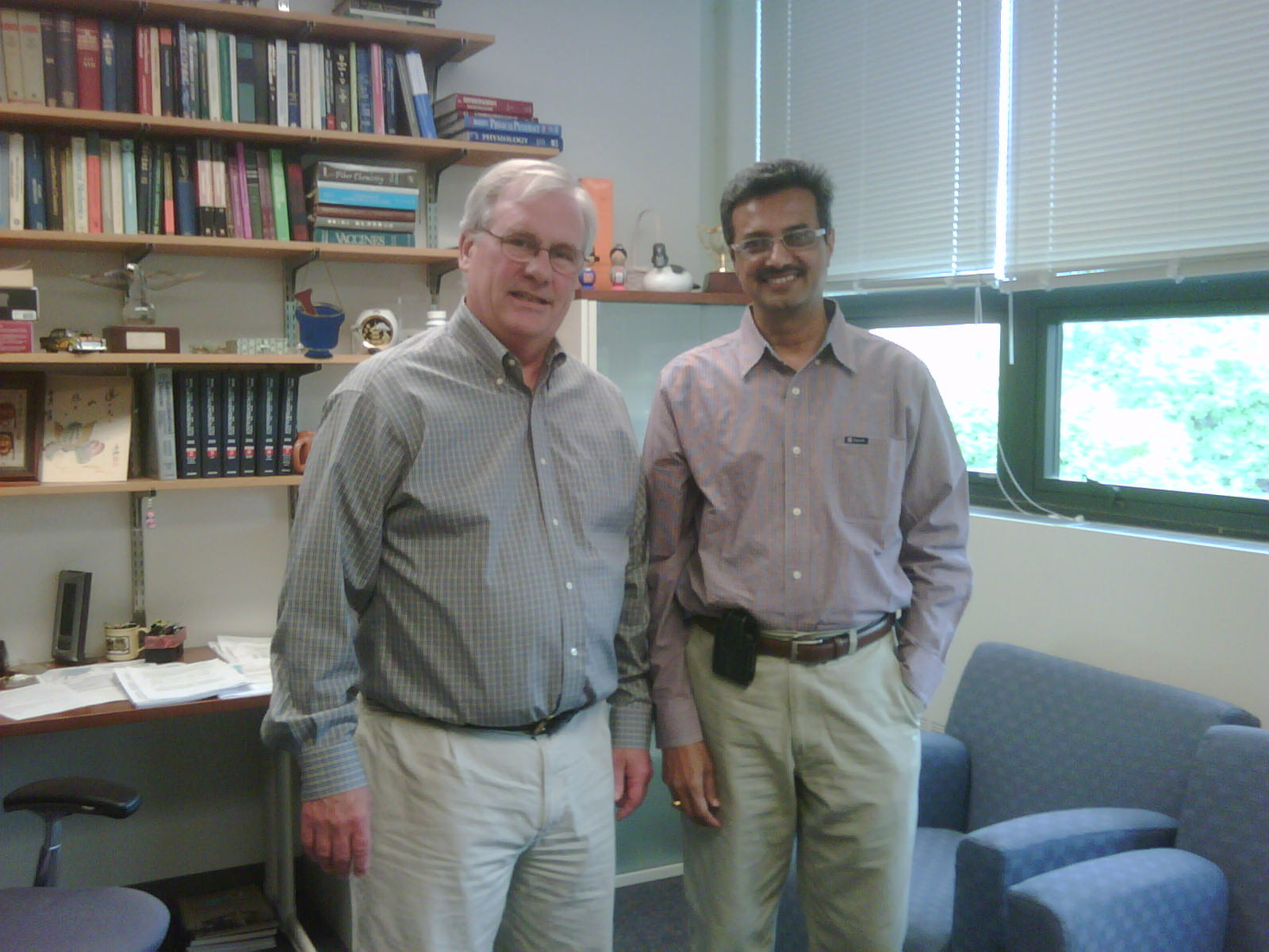 Two men standing side by side in an office. They are both wearing casual clothes and are standing in front of a bookshelf filled with books and other office supplies. The man on the left is older and is wearing a blue checkered shirt and beige pants while the man in the middle is younger. Both men are smiling and appear to be posing for the photo. There is a window with white blinds in the background and a blue armchair on the right side of the image. The room appears to be well-lit with natural light coming in from the window.