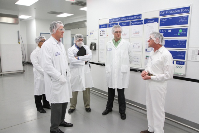 A group of six men in a laboratory setting. They are all wearing white lab coats and head caps and are standing in front of a whiteboard with blue and white posters on it. The posters appear to be related to a production board as there are several graphs and charts on the board. The men are engaged in a conversation with one man holding a clipboard and another holding a pen. They seem to be discussing something intently. The room has a high ceiling with fluorescent lights and there is a door on the left side of the image.