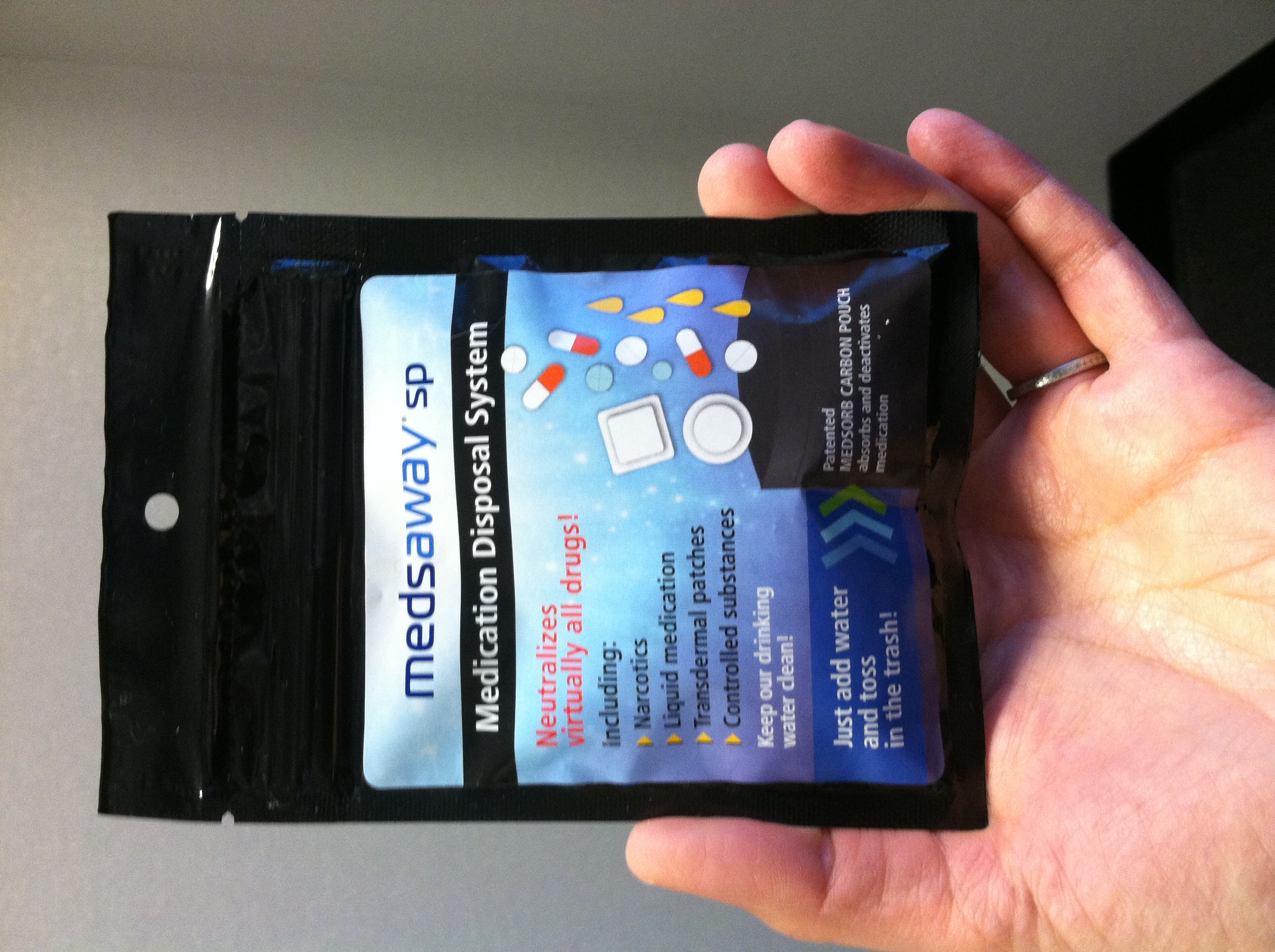 A hand holding a black packet labeled Medsaway SP Medication Disposal System. The packet is rectangular in shape and has a blue and white label on the front. The left side of the label has the text "Neutralizes virtually all drugs!" and includes a list of drugs. The label also reads "Keep our drinking water clean!" and "Just add water and toss in the trash!" On the right side of the label there is an illustration of various pills tablets liquids and patches falling into a black packet.