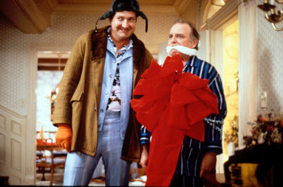 Two men standing in a room with a chandelier hanging from the ceiling. The man on the left is wearing a brown jacket blue jeans and a black hat with a feather on top. He is smiling and looking at the camera. On the right is an older man wearing a blue and white striped shirt and a red cape. He has a white mask covering his mouth and nose. The background shows a dining room with tables and chairs and a vase of flowers. The overall mood of the image is humorous and lighthearted.