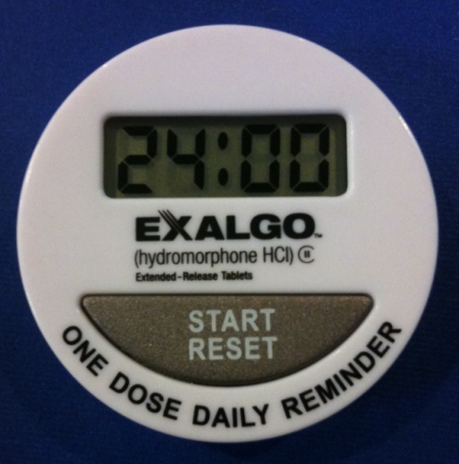 A close-up of a digital alarm clock. The clock is white in color and has a round shape. The top of the clock has a digital display that shows the time as 24:00. Below the display there is a text that reads "Exalgo (hydromorphone HCI) Extended-Release Tablets". Below the text it says "Start Reset" and "One Dose Daily Reminder". The clock appears to be mounted on a blue surface.