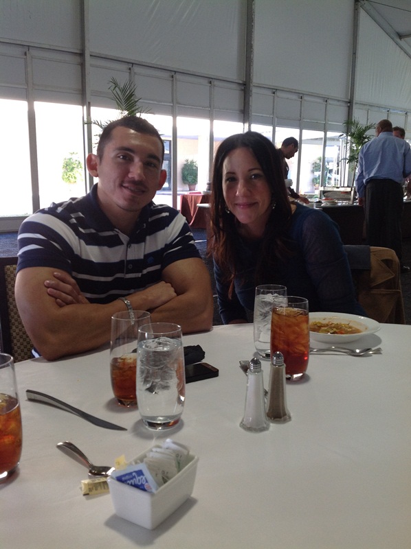 A man and a woman sitting at a table in a restaurant. They are both smiling and looking at the camera. The man is on the left side of the image and the woman on the right side. The table is covered with a white tablecloth and there are several glasses of drinks on it. There are also salt and pepper shakers on the table. In the background there are other people sitting at tables and a large window. The restaurant appears to be well-lit with natural light coming in from the windows.