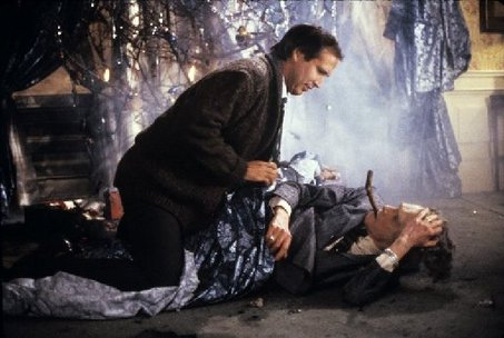 A scene from a movie or TV show. It shows a man kneeling on the ground next to a dead man lying on the floor. The man is wearing a dark suit and tie and appears to be in distress. He is holding a knife in his right hand and is looking down at the man with a concerned expression on his face. The background is dark and there is a hint of smoke or fog in the air. The overall mood of the image is tense and somber.