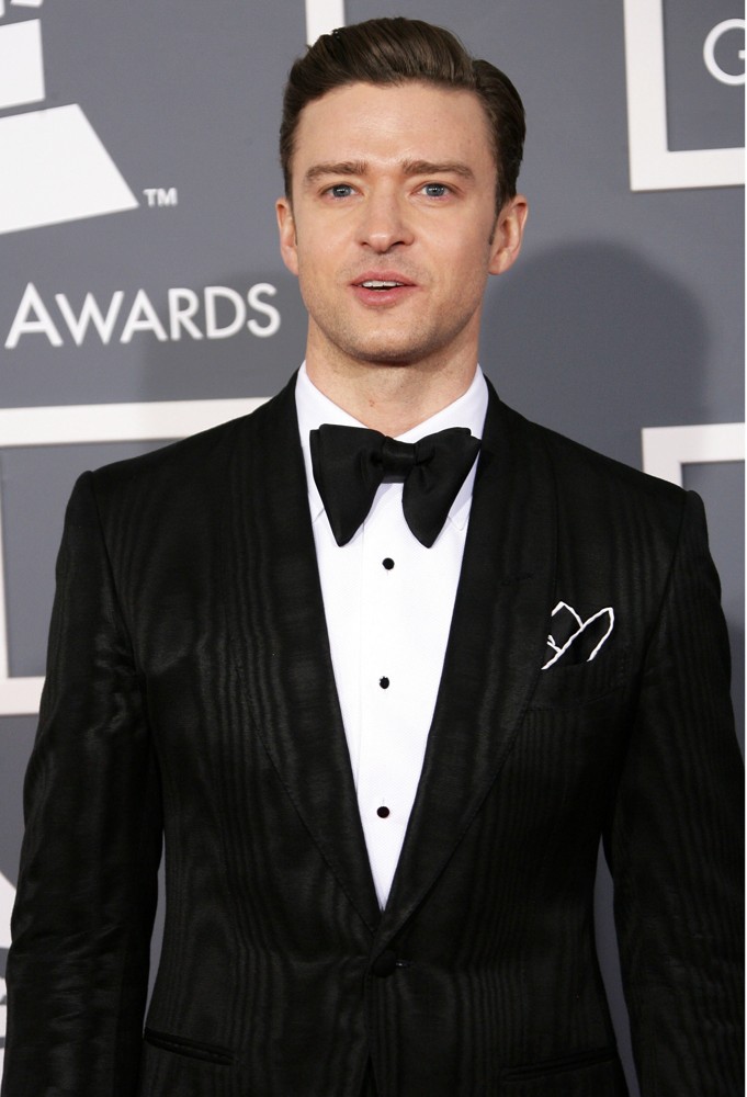 A portrait of a man posing on the red carpet at the Grammy Awards. He is wearing a black tuxedo with a white shirt and a black bow tie. The man has short dark hair styled in a neat slicked-back manner and is looking directly at the camera with a slight smile on his face. The background is a gray wall with the Grammy logo and the words "Grammy Awards" written in white.