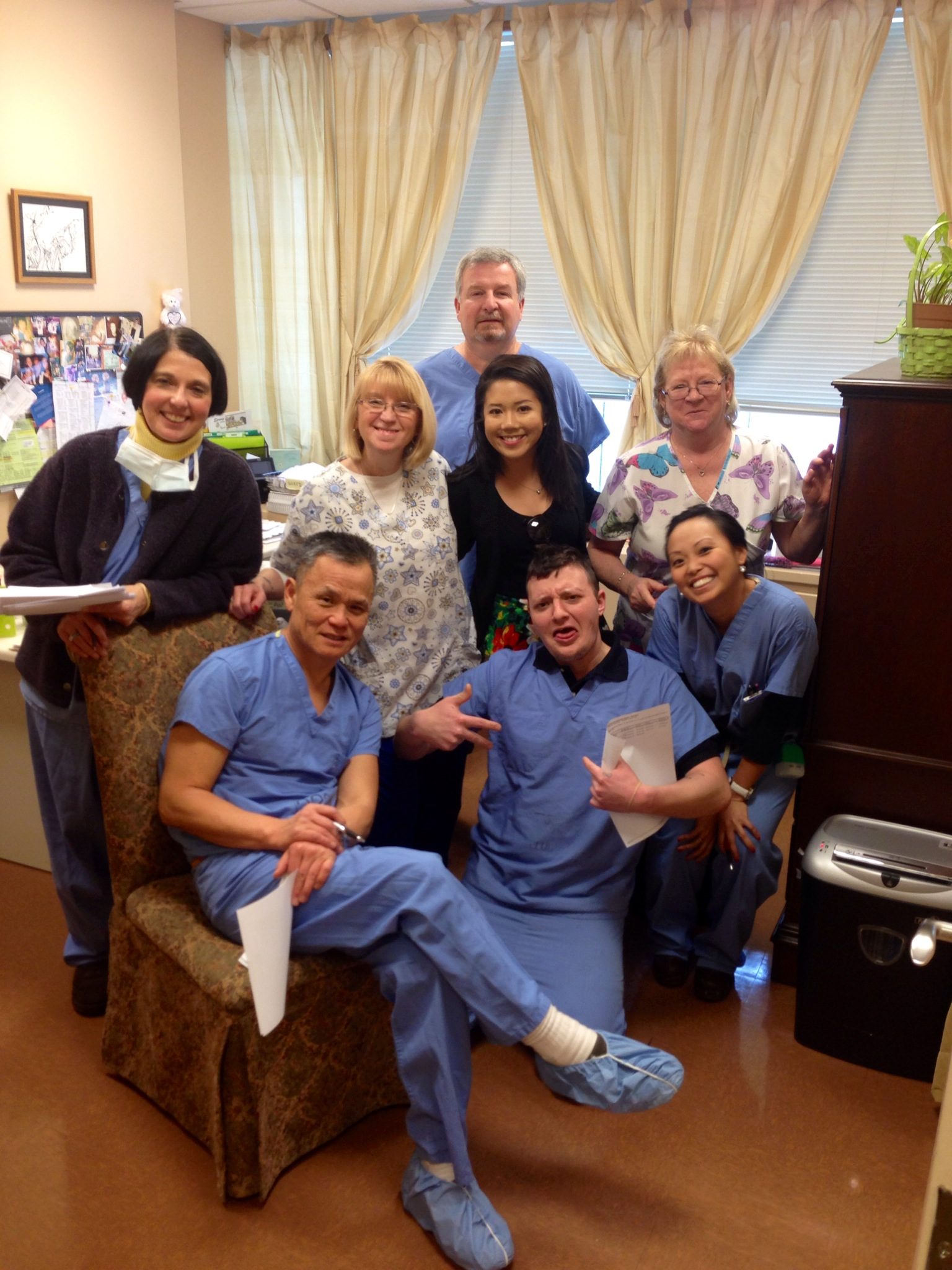 A group of nine people nine women and one man posing for a photo in a dental office. They are all wearing blue scrubs and are standing in front of a window with white curtains. The man in the center is sitting on a chair with his legs crossed and is holding a piece of paper in his hand. The other nine people are standing around him smiling and looking at the camera. On the left side of the image there is a woman wearing a face mask and holding a clipboard. In the background there are various dental equipment and a bulletin board with pictures and papers pinned to it. The overall mood of the photo is happy and relaxed.