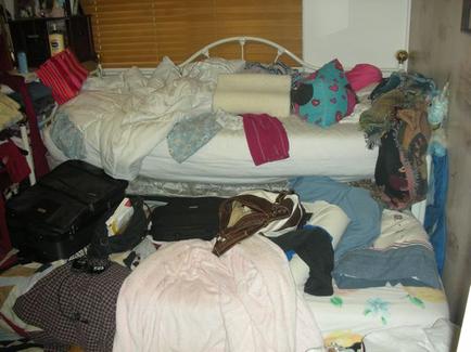 A messy bedroom with a white bed in the center. The bed is covered in a white comforter and there are several pillows and blankets scattered around it. On the left side of the bed there is a black suitcase and a black bag on the floor. The room appears to be cluttered with various items including clothes bags and other miscellaneous items. The walls are painted in a light color and the floor is covered with a patterned rug. There is a window with blinds in the background.