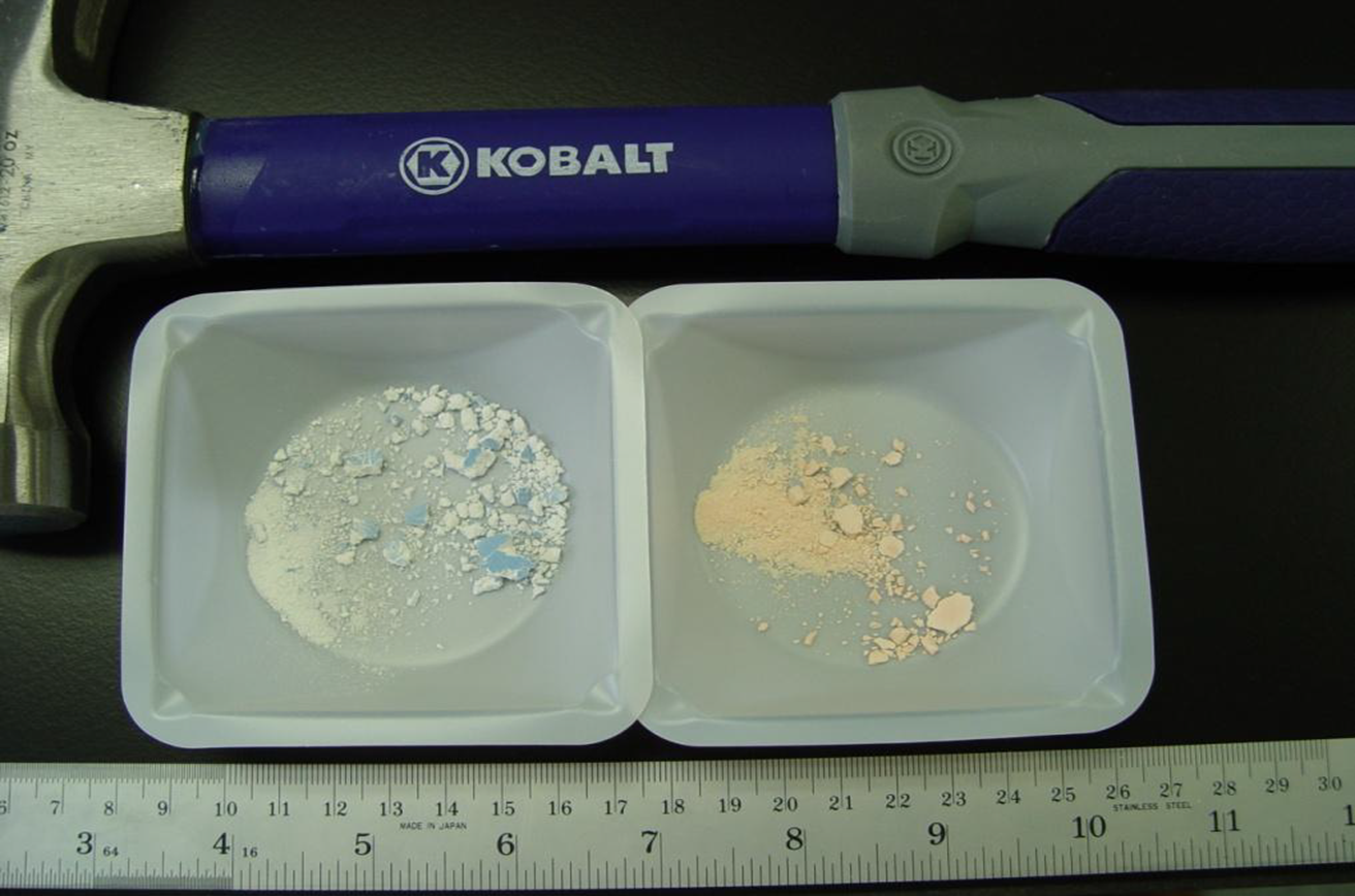 A Kobalt hammer with a blue handle and a silver head. The hammer is resting on a black surface with a ruler next to it. On the left side of the image there is a small white plastic container with two compartments. Inside the compartments there are two small pieces of white powder one of which appears to be a mixture of blue and white crystals. The powder is spilling out of the container onto the right side. The ruler is also visible in the image.