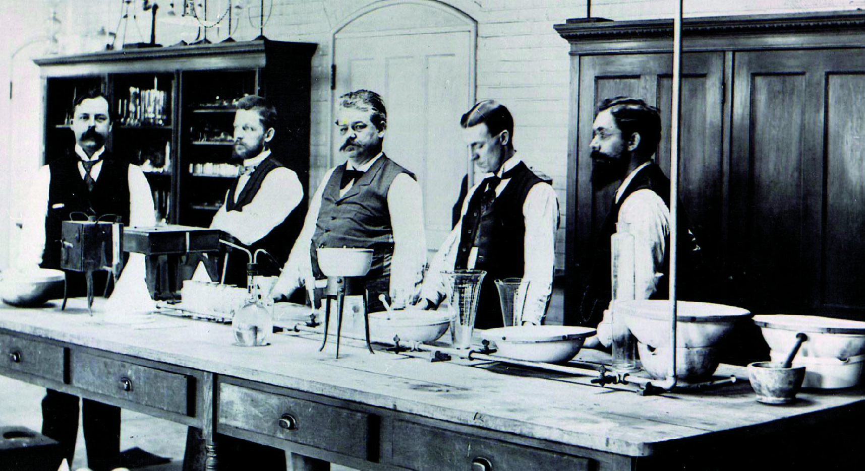 A group of six men standing around a long wooden table in a laboratory. They are all wearing vests and appear to be in the middle of a scientific experiment. The table is covered with various scientific equipment including a microscope a beaker a flask and several bowls and containers. The men are standing in front of a bookshelf and there is a door in the background. The image is in black and white and appears to be from the early 20th century.