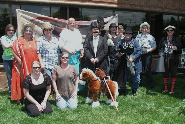 A group of people posing for a photo in front of a brick building. There are nine people in the photo all dressed up in various costumes and hats. The group is standing on a grassy lawn and there is a large American flag in the background. In the center of the group there is an older man wearing a black suit and a top hat holding a cane. To his left there are two women wearing colorful dresses and hats one wearing a red dress and the other wearing a blue dress. To the right there appears to be a man dressed as a pirate with a sword and shield. In front of him is a stuffed toy horse. Everyone is smiling and looking at the camera.