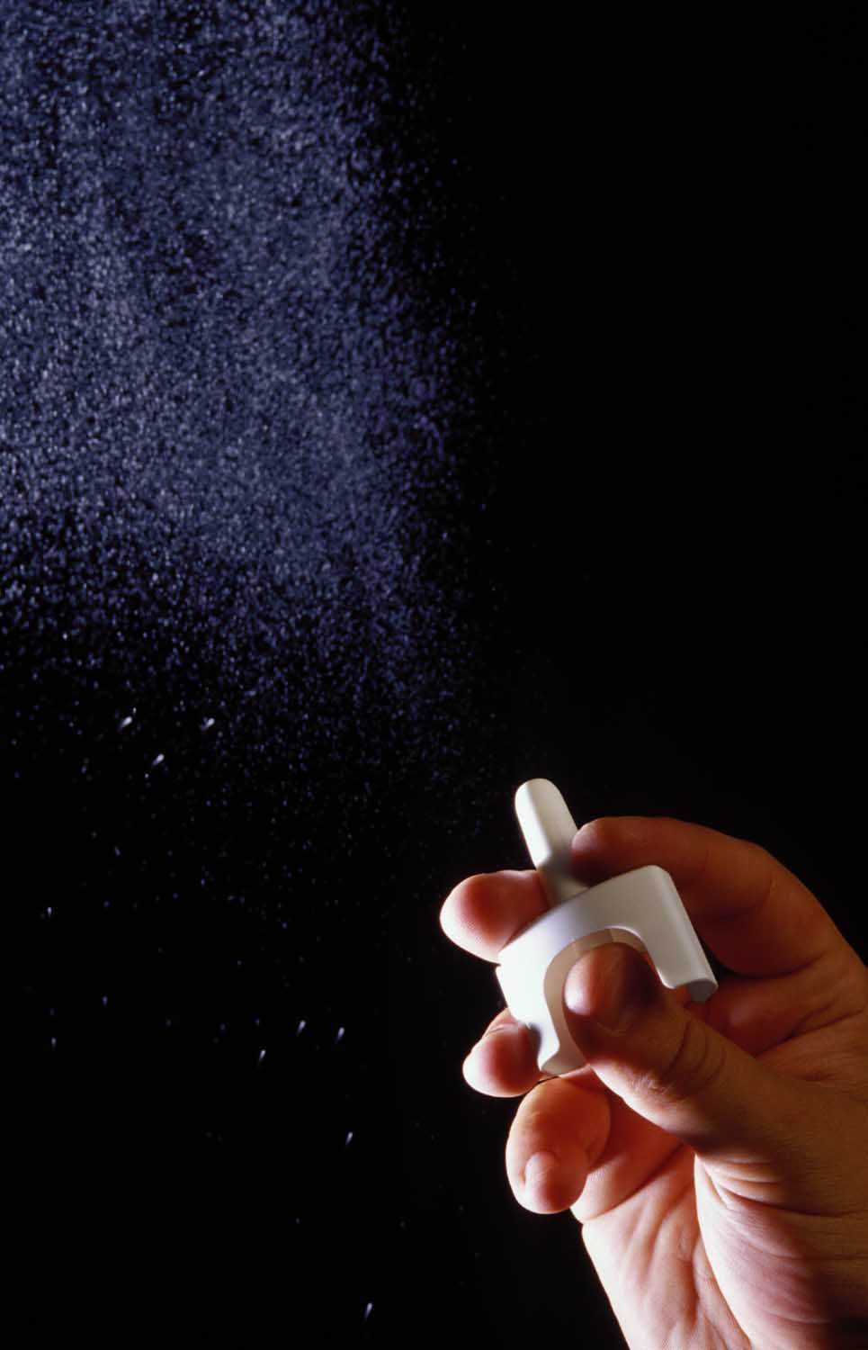 A hand holding a small white object which appears to be a sprinkler against a black background. The sprinkler is spraying water onto the ground creating a cloud-like effect. The water droplets are visible in the air and the hand is holding the object with one hand. The image is taken from a low angle so the focus is on the hand and the sprinkler. The overall mood of the image is dark and mysterious.