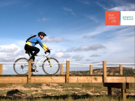 A person riding a mountain bike on a wooden bridge. The person is wearing a blue and yellow jersey black pants and a black helmet. The bike is white and black and the rider is in the middle of a jump. The bridge is made of wooden planks and has a railing on one side. The background shows a clear blue sky with some clouds. The image appears to be taken on a sunny day.