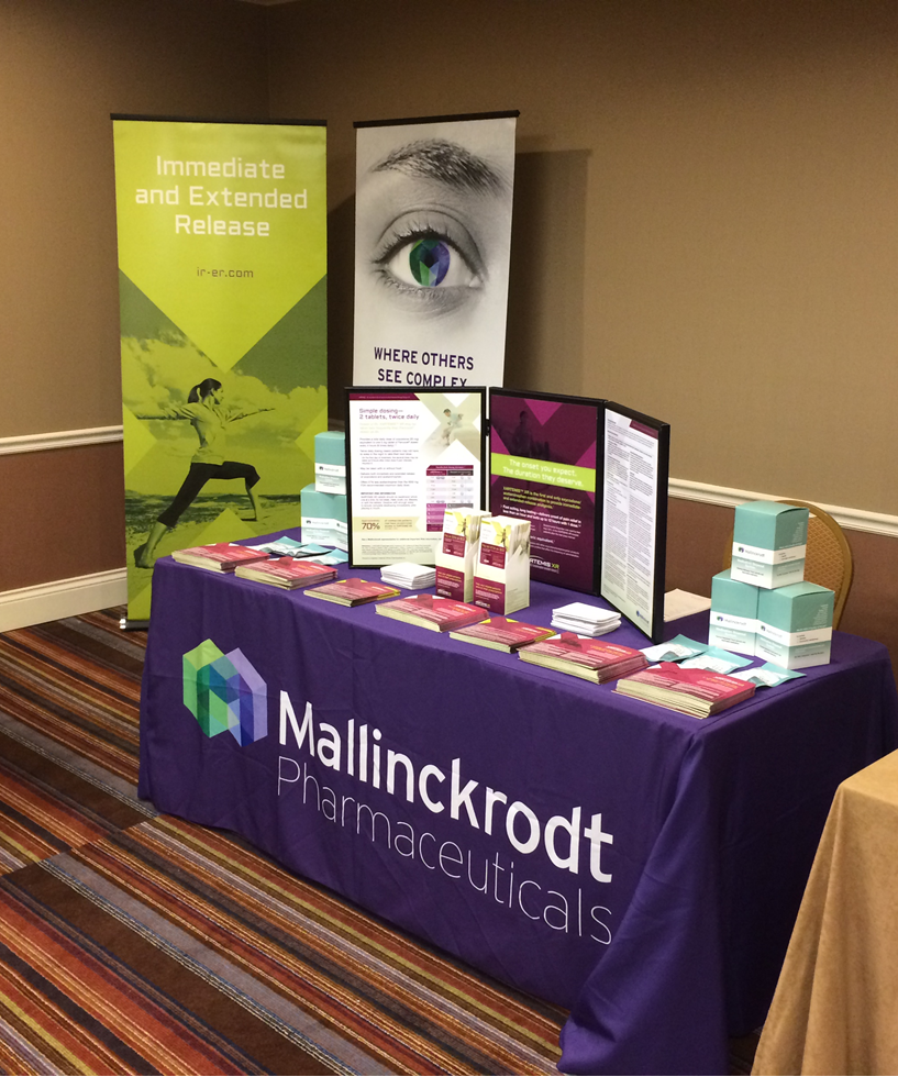 A table with a purple tablecloth and a banner that reads "Mallinckrodt Pharmaceuticals". On the table there are several books and pamphlets arranged in a neat and organized manner. The table is set up in a room with beige walls and a carpeted floor. On the left side of the image there is a banner with an image of an eye and the text "Immediate and Extended Release". On top of the banner there appears to be a sign that says "Where others see you". The table also has a logo of the company on it.