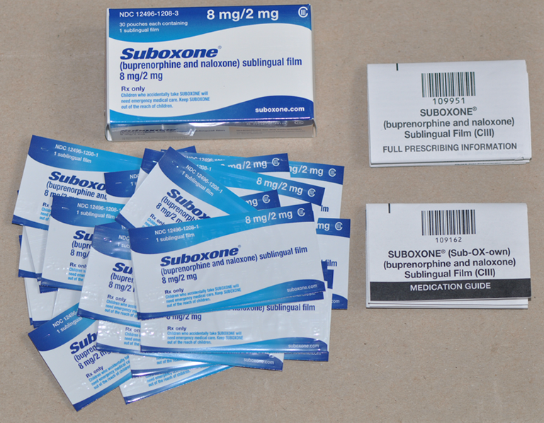 A collection of medical supplies including a box of Suboxone a medication guide and a full-prescription information card. The box is white with blue text that reads "Suboxone 8 mg/2 mg" and has a barcode on the right side. There are also several small packets of the medication which are blue and white in color. The packets are arranged in a scattered manner with some overlapping each other. The background is a beige-colored surface.