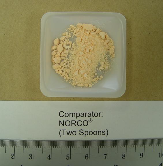 A small square plastic container with a white label on it. The label reads "Comparator: NORCO (Two Spoons)". Inside the container there is a pile of small light-colored granules or granules. The granules appear to be finely ground and have a rough texture. The container is placed on a beige-colored surface.