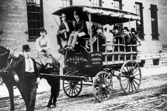 A black and white photograph of a group of men sitting on top of a horse-drawn carriage. The carriage is on a street with a brick building in the background. The men are dressed in formal attire with some wearing hats and others wearing suits. The man on the left is standing next to the carriage while the man in the center is sitting on the back of the carriage. There are several other men sitting in the carriage and one man standing on the right side of the image. All of them are looking at the camera and appear to be posing for the photo.<br /><br />The carriage has a sign on the side that reads "Mallory's Chemical Works." The carriage appears to be old and weathered with peeling paint and rust visible on the sides. The horses are harnessed to the cart and are standing on a cobblestone street. The photograph appears to have been taken in the early 20th century.