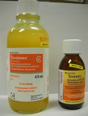 Two bottles of Tussionex products. The bottle on the left is yellow in color and has a white label with the brand name "Tassoni" written in bold black letters. The label also has the product name "473 ml" written on it in smaller black letters indicating that it is a 12-hour dosing solution for children under 6 years of age. Next to the bottle there is a smaller bottle with a white cap. Both bottles are placed on a white surface. The background is blurred but it appears to be a laboratory setting.