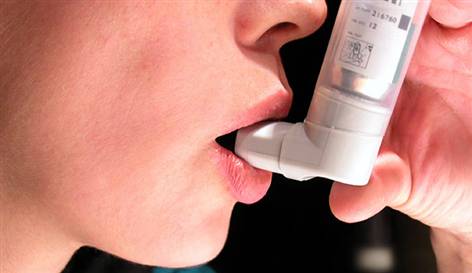 A close-up of a person's face with their mouth open as they use an asthma inhaler. The inhaler is white in color and has a label on it with a barcode and some text. The person's hand is visible on the right side of the image holding the inhaler and using it to inhale the air. The background is blurred so the focus is on the person and the device.