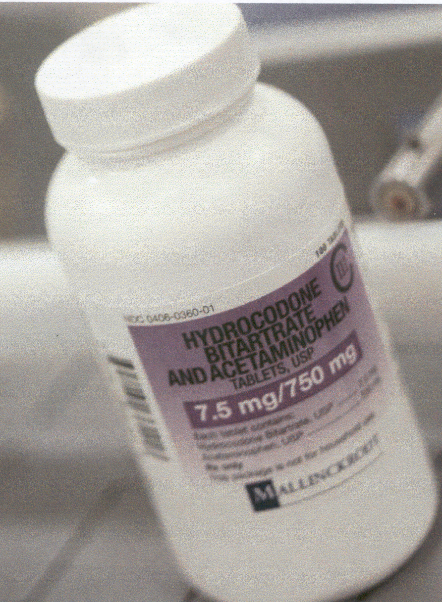 A close-up of a white plastic bottle with a purple label. The label has the brand name "Hydrocodone" written in bold black letters at the top followed by the product name "Bitartrate and Acetaminophen" in smaller black letters. Below that there is a description of the product which states that it is 7.5 mg/750 mg. The bottle has a white cap and is sitting on a white surface. The background is blurred but it appears to be a kitchen countertop.