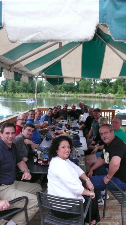 A group of people sitting at a long table on a wooden deck overlooking a lake. The table is covered with a green and white striped awning and there are several bottles of beer and other drinks on the table. The people are smiling and appear to be enjoying themselves. In the background there are trees and a boat on the water. The sky is blue and the overall atmosphere is relaxed and casual.