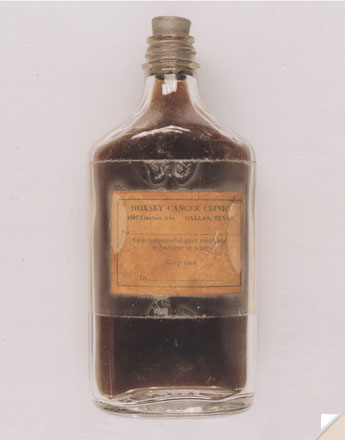 Of a small glass bottle with a metal stopper. The bottle is filled with a dark brown liquid. The label on the bottle is orange and has the words "Hoxsey Cancer Clinic" written in bold black letters. The bottle appears to be very old.