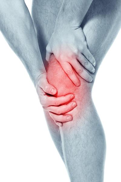 A close-up of a person's lower body specifically their lower leg. The person is holding their knee in pain with their hands gently cradling it. The knee appears to be swollen and inflamed with red spots covering the area. The skin around the knee is smooth and shiny and the person's legs are slightly bent at the knees. The background is white making the red spots stand out.