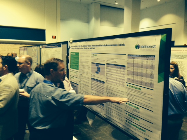 A group of people gathered around a large poster board in a conference room. The poster board is white with green and blue text and images on it. The man in the center of the image is pointing at the poster board with his right hand and appears to be explaining something to the group. He is wearing a blue shirt and is standing in front of it. There are other people in the background some of whom are looking at the board and others are engaged in conversation. The room has a high ceiling with white walls and a large window on the left side.