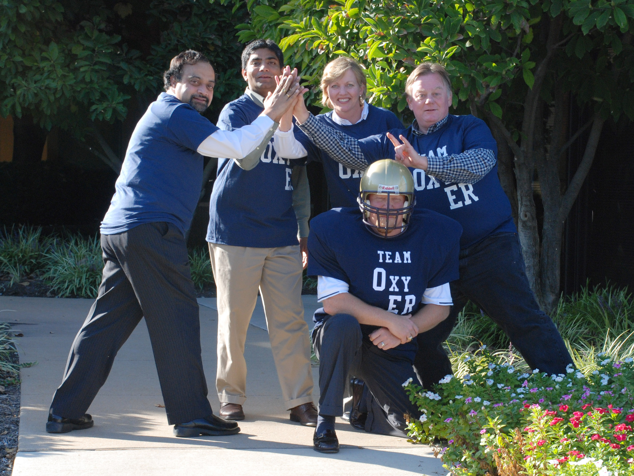 A group of five people four men and one woman posing for a photo in a garden. They are all wearing blue t-shirts with the words "Team Oxygen" printed on them. The man in the center is wearing a football helmet and is kneeling on the ground. The other three people are standing around him clapping their hands in a celebratory manner. The garden has a variety of plants and flowers and there are trees and bushes in the background. It appears to be a sunny day.