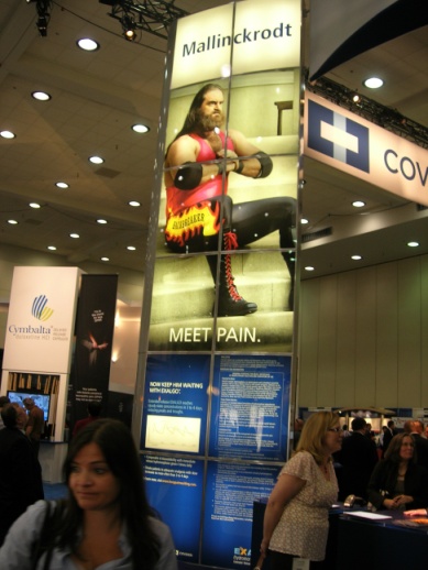 A large banner stand in a trade show booth. The banner has a picture of a man with long hair and a beard wearing a red tank top and black pants. He is holding a pair of boxing gloves and appears to be in the middle of a boxing match. The text on the banner reads "Mallinckrodt" and "Meet Pain." Below the banner there is a blue banner with the company's logo and contact information. In the background there are other booths and people walking around the booth.