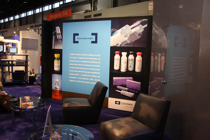 A booth at an exhibition or trade show. The booth has a blue background with white text and images. On the left side of the booth there is a large banner with the company's logo and contact information. <br /><br />In front of the banner there are two black leather armchairs and a round glass coffee table. The armchairs have a modern design with a curved backrest and armrests. The coffee table has a glass top and is placed in the center of the room.<br /><br />The booth also has a display of different types of hair care products including shampoo conditioner and conditioner bottles. There are also images of a hair dryer and a hair straightener on the banner. In the background we can see other booths and equipment suggesting that the booth is located in a large industrial space with high ceilings and large windows.