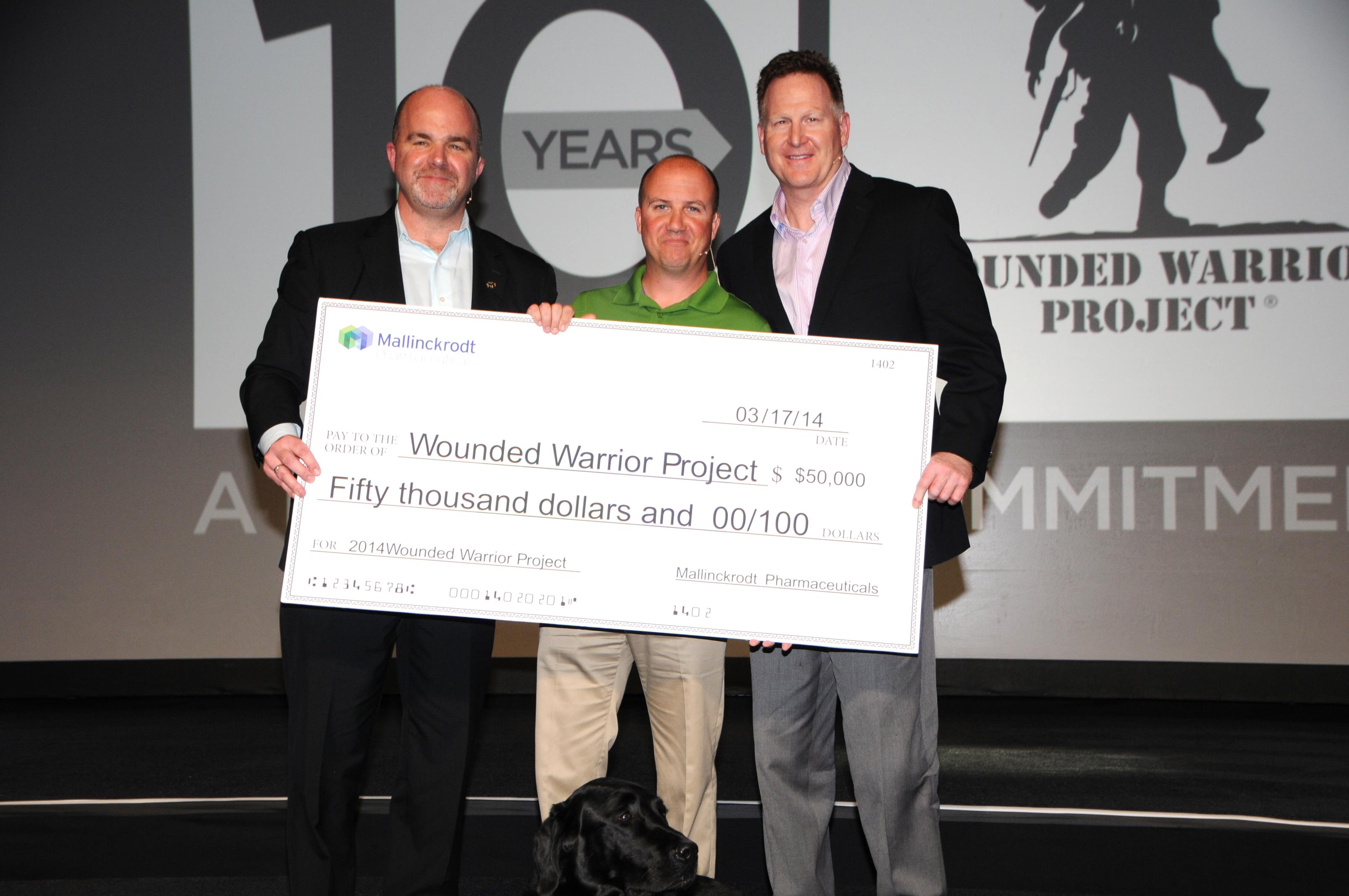 Three men standing on a stage with a large check in front of them. The check is for $50000 and is being presented to the wounded warrior project. The man in the middle is wearing a green shirt and is holding the check with both hands. The two men on either side of him are wearing suits and are smiling at the camera. In the background there is a large screen with the logo of the project and the words "Wounded Warrior Project" and "Commitment" on it. A black dog is sitting on the stage next to the check.