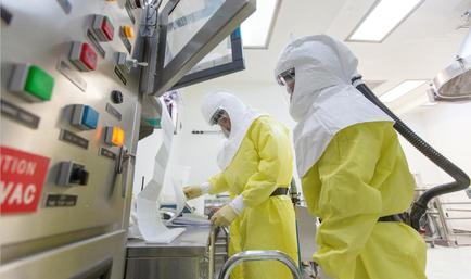 Two people wearing yellow protective suits and helmets working in a laboratory. They are standing in front of a control panel with various buttons and switches. The person on the left is holding a piece of paper and appears to be working on a machine while the person in the middle is using a vacuum cleaner. The control panel has a red sign that reads "VAC" and there is a large screen above it. The background shows other laboratory equipment and equipment in the background. The overall atmosphere of the image is clean and sterile.