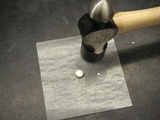 A hammer with a wooden handle and a metal head. The hammer is resting on a square piece of what appears to be parchment paper. On the left of the hammer there is a small white pill. The background is a black countertop.