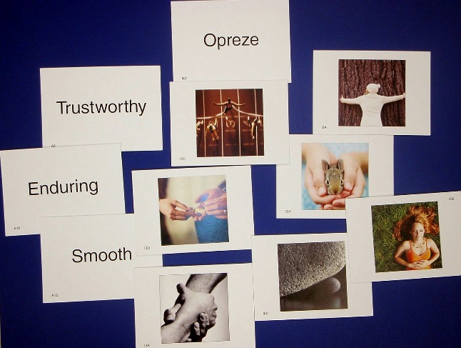 A photograph of eleven index cards arranged on a blue surface. There are four index cards arranged mostly on the left that have the words "Opreze" "Trustworthy" "Enduring" and "Smooth". On the right side the remaining seven index cards all have photographs that feature hands in some way all with different people doing different things. Some examples include a hand holding a rock a woman hugging a tree runners on a track and hands holding a chipmunk.