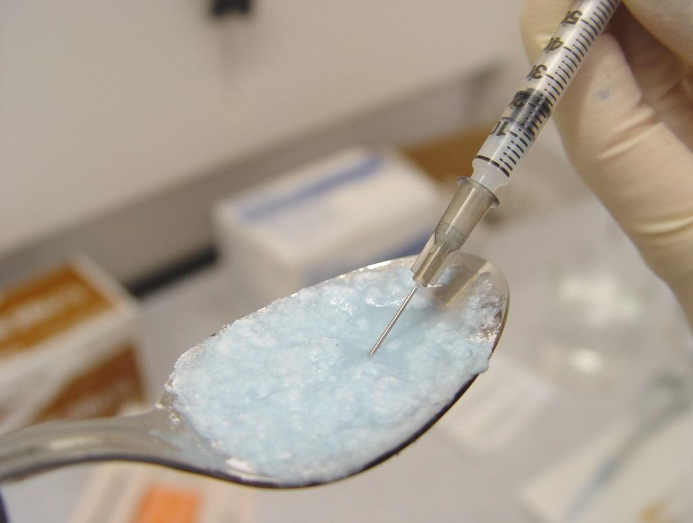 A spoon filled with a light blue substance likely heroin or another drug and a hand holding a syringe. The syringe drawing up the substance.  The hand is wearing a white safety glove. The background is blurred but it appears to be  laboratory setting.