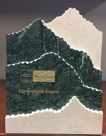 Of a cornerstone award. The award is made of a dark green marble-like material with a white base. The top half of the award is shaped like a mountain peak while the bottom half is a lighter shade of green. On the base there is a gold-colored plaque with the words "Cornerstone Award" written in black. The plaque appears to be made of ceramic or porcelain.