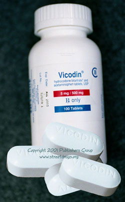 A white plastic bottle with a white cap and a blue label. The label has the brand name "Vicodin" written in bold black letters at the top followed by the product name "5 mg/500 mg" in smaller black letters. Below that there is a description of the product which states that it is only 100 tablets. The bottle is placed on a dark green surface and there are three white pills scattered around it. The pills appear to be of different sizes and shapes.