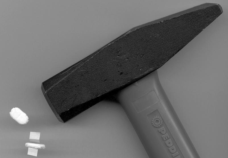 A black and white photograph of a hammer. The hammer has a long pointed head with a sharp pointed tip. The handle of the hammer is grey and has the word "PEDDI" written on it in white letters. On the left side of the image there are two small white objects one of which appears to be a piece of paper or fabric. The background is plain and the focus is on the hammer and the objects.