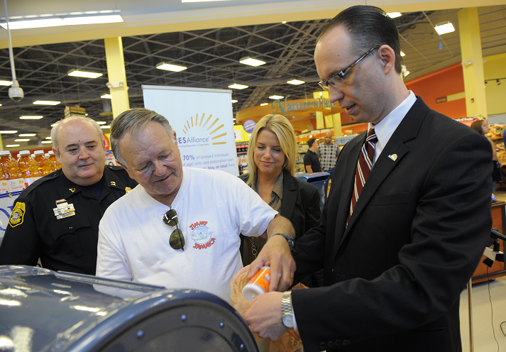 A group of people in a store. There are three people in the foreground two men and a woman. The man on the left is wearing a white t-shirt with a red logo on it and sunglasses. He is holding a can of orange juice and appears to be pouring it into a black container. The woman on the right is standing next to him wearing a black suit and glasses. In the background there are shelves stocked with orange juice bottles and a banner with the store's logo. The store appears to have a high ceiling with overhead lights.