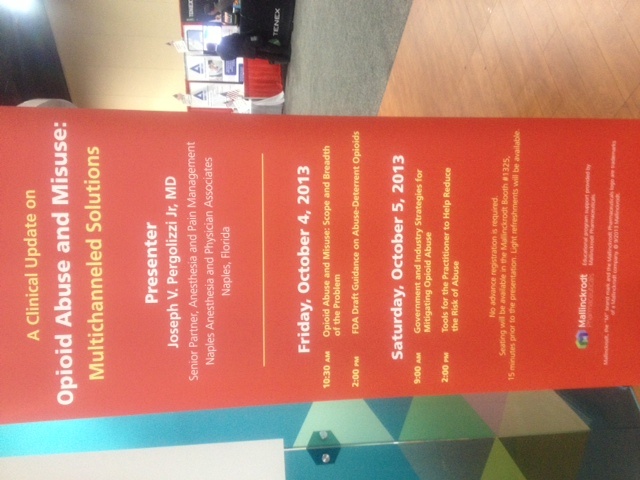 A photograph of a red banner with white text. The banner is hanging on a wooden floor in a room with a white wall and a wooden door in the background. The text on the banner reads "Opioid Abuse and Misuse: A Clinical Update on Multichannel Solutions". Below the text there is a list of events taking place on Friday October 4 2013. The events are listed in a table format with columns for each event. The date and time of the event is also visible on the right side of the banner.