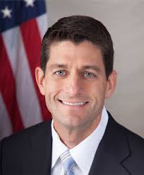 A professional headshot of a man. He is wearing a dark suit and a light blue tie. He has short dark hair and is smiling at the camera. The background is blurred but it appears to be an American flag. The man looks confident and professional.